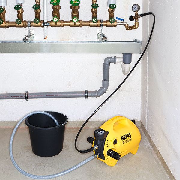 A yellow and black REMS Pro 3000 water pressure washer connected to a gray hose and a black bucket, set against a white wall with a water supply pipe in the background.